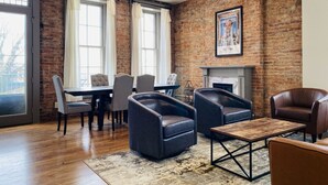 A modern living room with a brick wall, a fireplace, leather armchairs, a dining table with chairs, and large windows. - 4 Bed, 3 Bath Loft in Downtown Louisville! Amazing River Views! Private Deck! (Louisville)