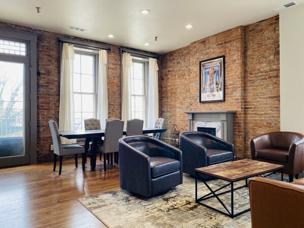 A modern living room with a brick wall, a fireplace, leather armchairs, a dining table with chairs, and large windows. - 4 Bed, 3 Bath Loft in Downtown Louisville! Amazing River Views! Private Deck! (Louisville)