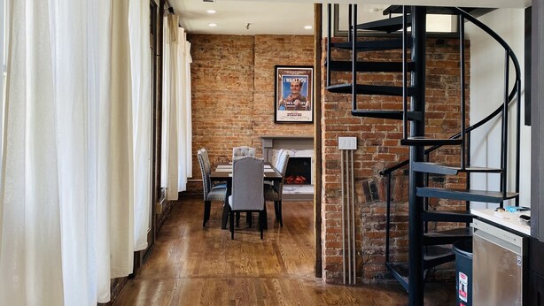 A modern interior with a wooden floor, a dining area with chairs and a table, a staircase with a metal railing, and a brick wall. - 4 Bed, 3 Bath Loft in Downtown Louisville! Amazing River Views! Private Deck! (Louisville)