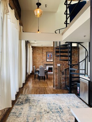 Un intérieur moderne avec un plancher en bois, un coin repas comprenant des chaises et une table, un escalier avec une rampe en métal et un mur en briques.