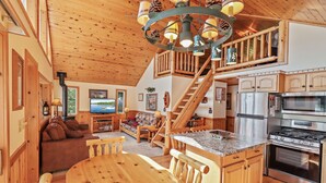 A spacious cabin interior with a wooden dining area, a cozy living room with a sofa and fireplace, a kitchen with modern appliances, and a staircase connecting the two levels.