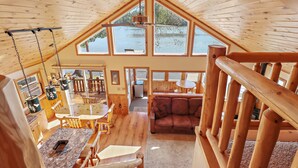 A spacious cabin with a high ceiling, wooden interior, a dining area, a seating area, and large windows overlooking a body of water.