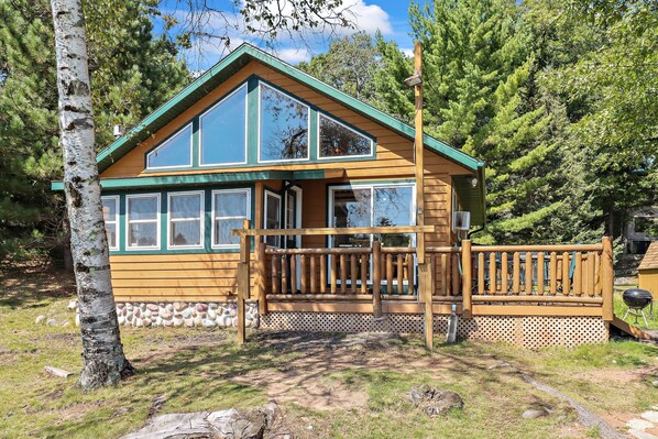 A wooden cabin with large windows and a porch, surrounded by trees and a stone wall. - Stepaway Vacation Rentals - Carefree Creek Cabin (Saint Germain)