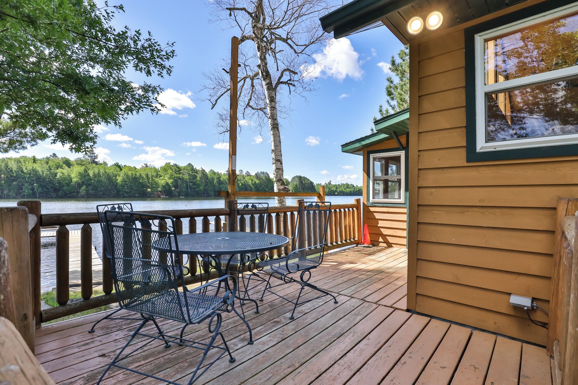 A wooden deck with metal chairs and a table, overlooking a lake with trees in the background.