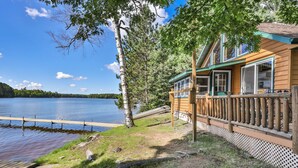 A cabin by a lake with a dock, surrounded by trees and a wooden deck.