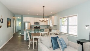 A spacious living room with a sofa, a dining area with chairs, a kitchen with a central island, and a large window.