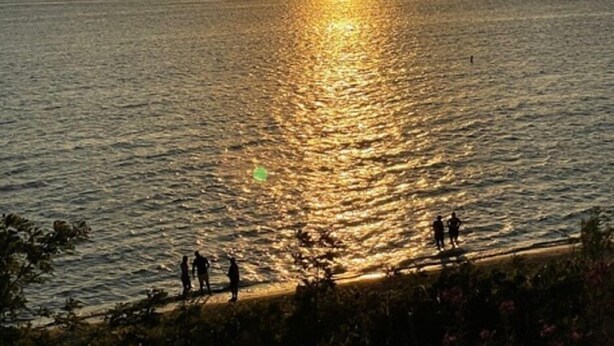 A sunset over a calm sea with a few people on the shore.