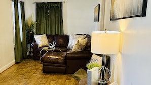 A living room with a brown leather sofa, a green armchair, a wooden floor, a lamp, and a painting on the wall.