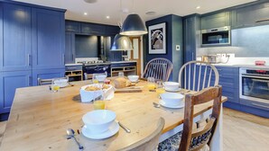 A modern kitchen with blue cabinets, a wooden dining table set with breakfast items, and a large pendant light. - Anchor Light Cottage (Faversham)