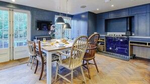 A modern kitchen with blue cabinets, a dining table with chairs, and a large window. - Anchor Light Cottage (Faversham)