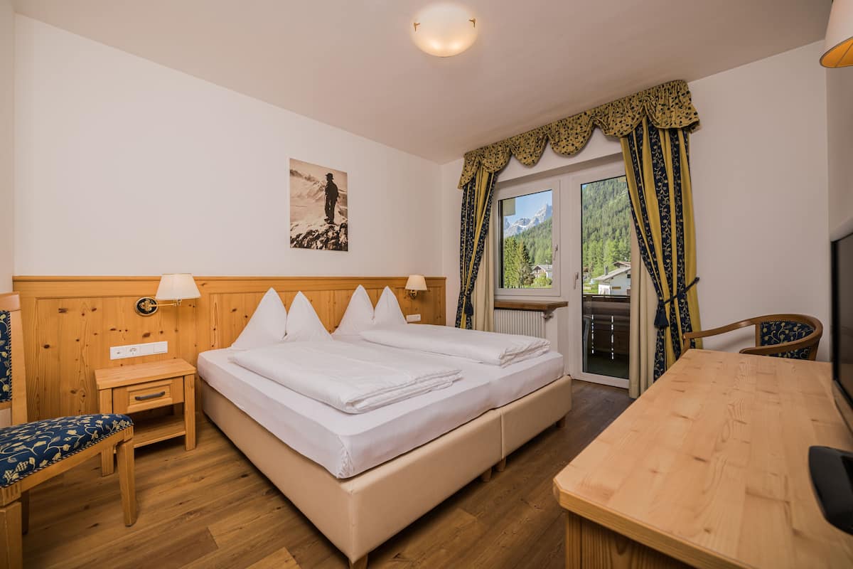 Ein Hotelzimmer mit einem hölzernen Bett, einem kleinen Nachttisch, einem Stuhl, einem Holztisch und Bergblick durch das Fenster.