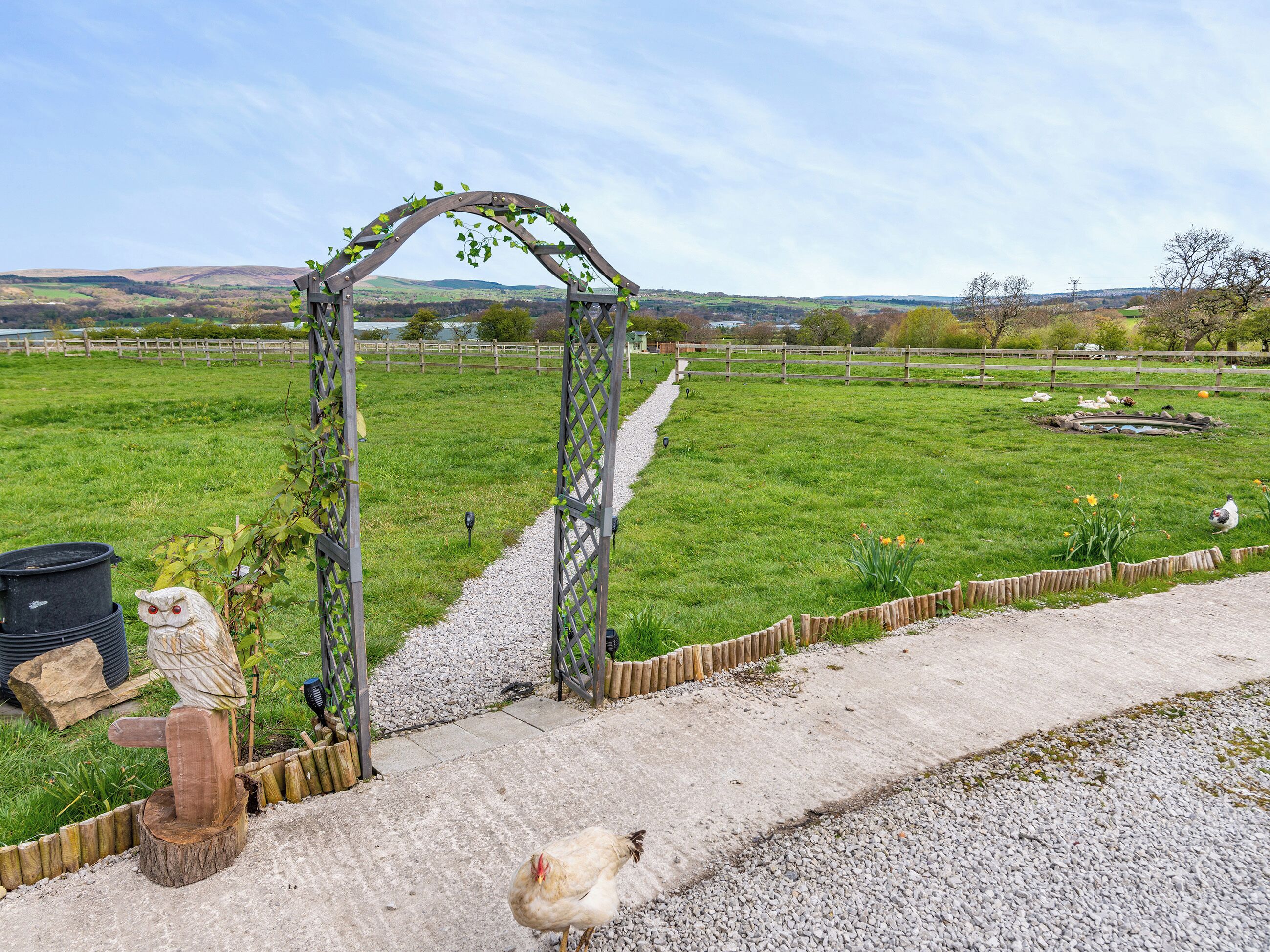 A rural landscape with a wooden archway, a gravel path, and a fenced field.