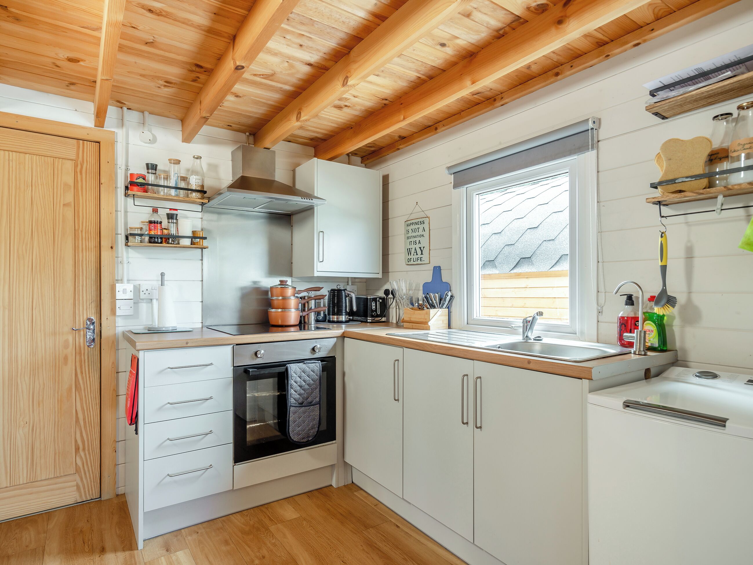 Una cocina compacta con vigas de madera, armarios blancos, horno integrado y una ventana con persianas.