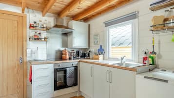 Una cocina compacta con vigas de madera, armarios blancos, horno integrado y una ventana con persianas.