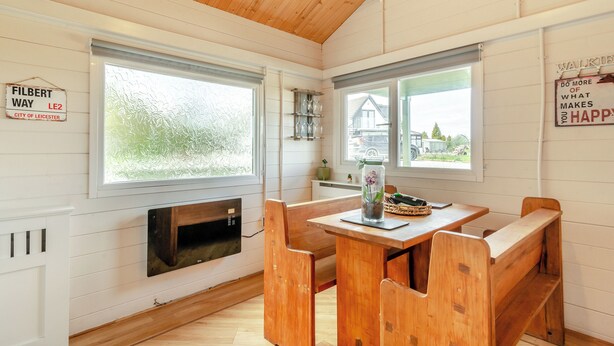 A cozy room with wooden walls and a sloped ceiling, featuring a dining table with benches, a fireplace, and large windows offering views of the outside. - 2 bedroom accommodation in Altham (Altham)