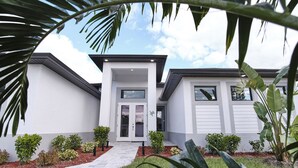 A modern two-story house with a white exterior, a front entrance with glass doors, and a driveway leading to a garage. - Waterfront home with pool and spa close to shopping (Cape Coral)