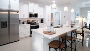 A modern kitchen with white cabinetry, stainless steel appliances, a central island with bar stools, and pendant lighting. - Waterfront home with pool and spa close to shopping (Cape Coral)