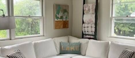 A corner living room with a white sectional sofa, a black coffee table, and a window with a view of trees.