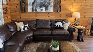 A cozy living room with a brown leather sofa, a large abstract painting, a wooden wall, and a small side table with a lamp. - Bear Cabin - Close to Magic Springs, Off-Road Park (Hot Springs)