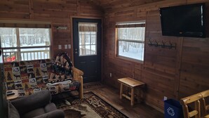 Un interior de cabaña acogedor, con detalles de madera, un sofá con colcha, un banco de madera pequeño, un televisor y una puerta con un cartel.