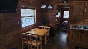 Interior de una cabaña con una mesa de comedor de madera, bancos, una cocina con un fregadero y una ventana con vistas a árboles.