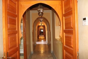 A wooden hallway with an ornate chandelier, leading to a room with a bed and a nightstand.