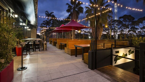An outdoor seating area with wooden benches and tables, string lights, and palm trees. - Nightcap at the Ranch Hotel (Sydney)