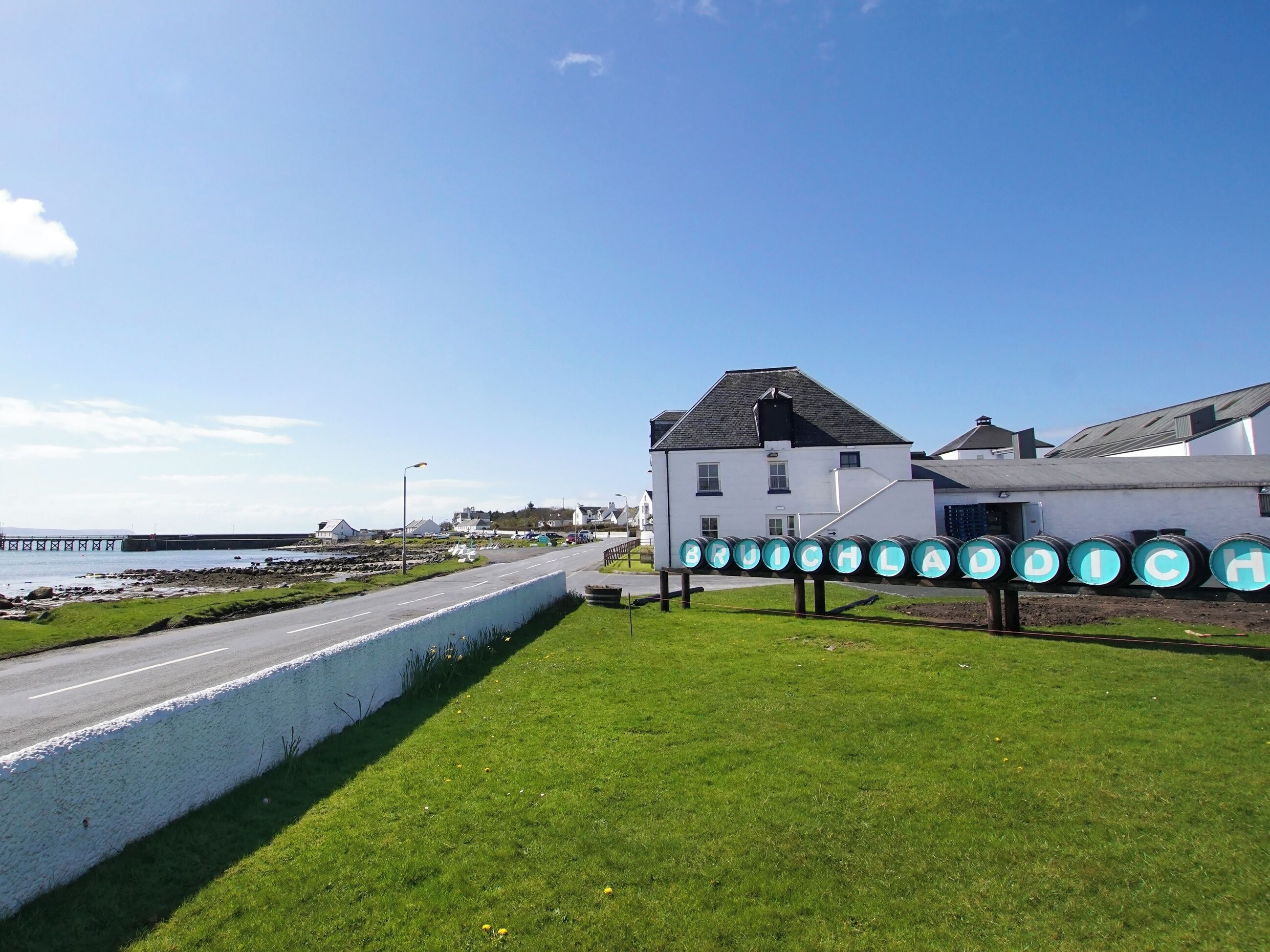 A coastal road with a white building and a row of barrels labeled "BRUICHALDA".