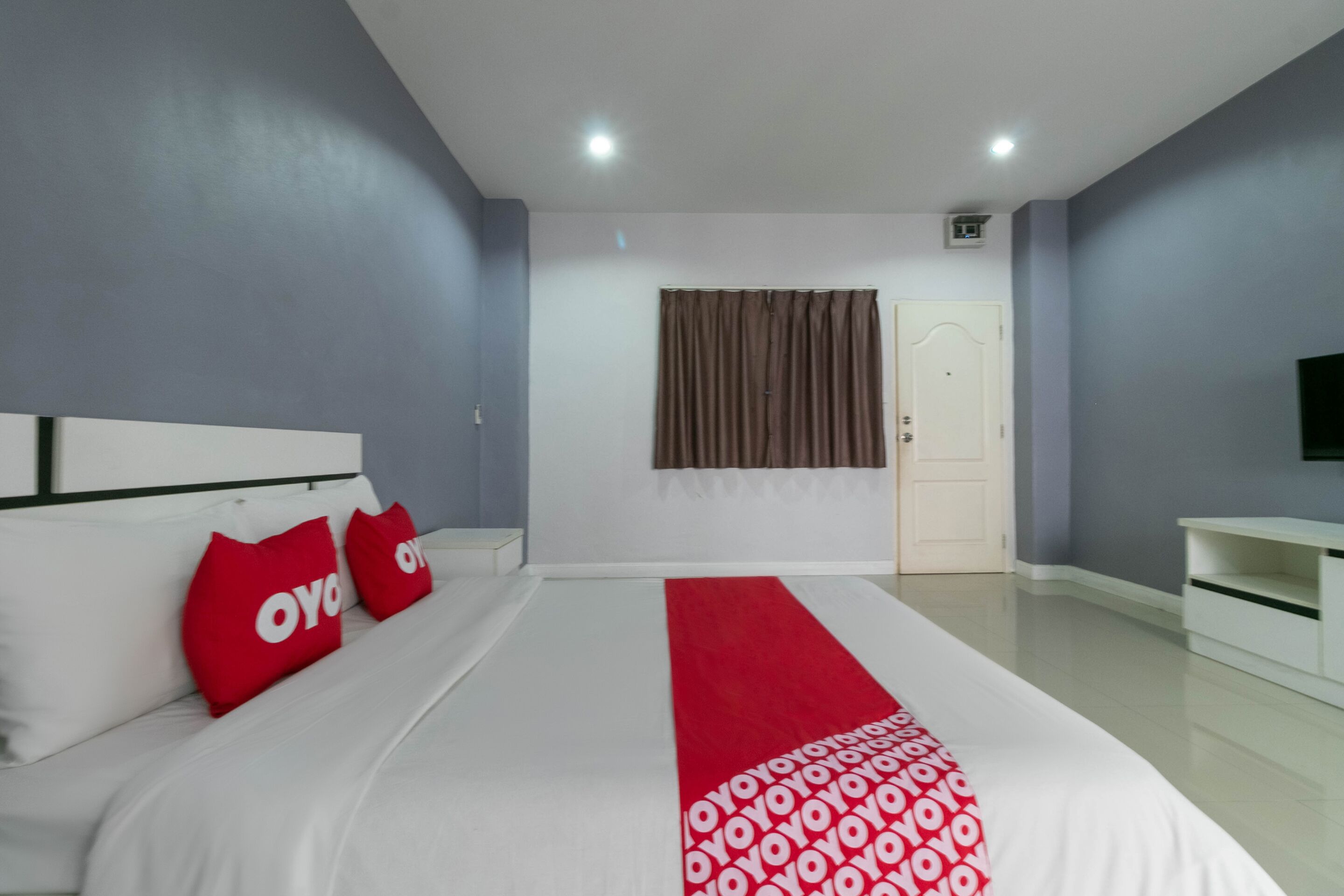 Una habitación de hotel con una cama grande, un corredor rojo y blanco estampado, un cabecero blanco y un televisor sobre un mueble.