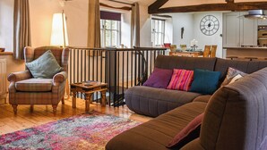 Un salon chaleureux avec un canapé, un fauteuil et une petite table. On y voit une fenêtre avec des rideaux, un escalier et, au fond, un coin cuisine.