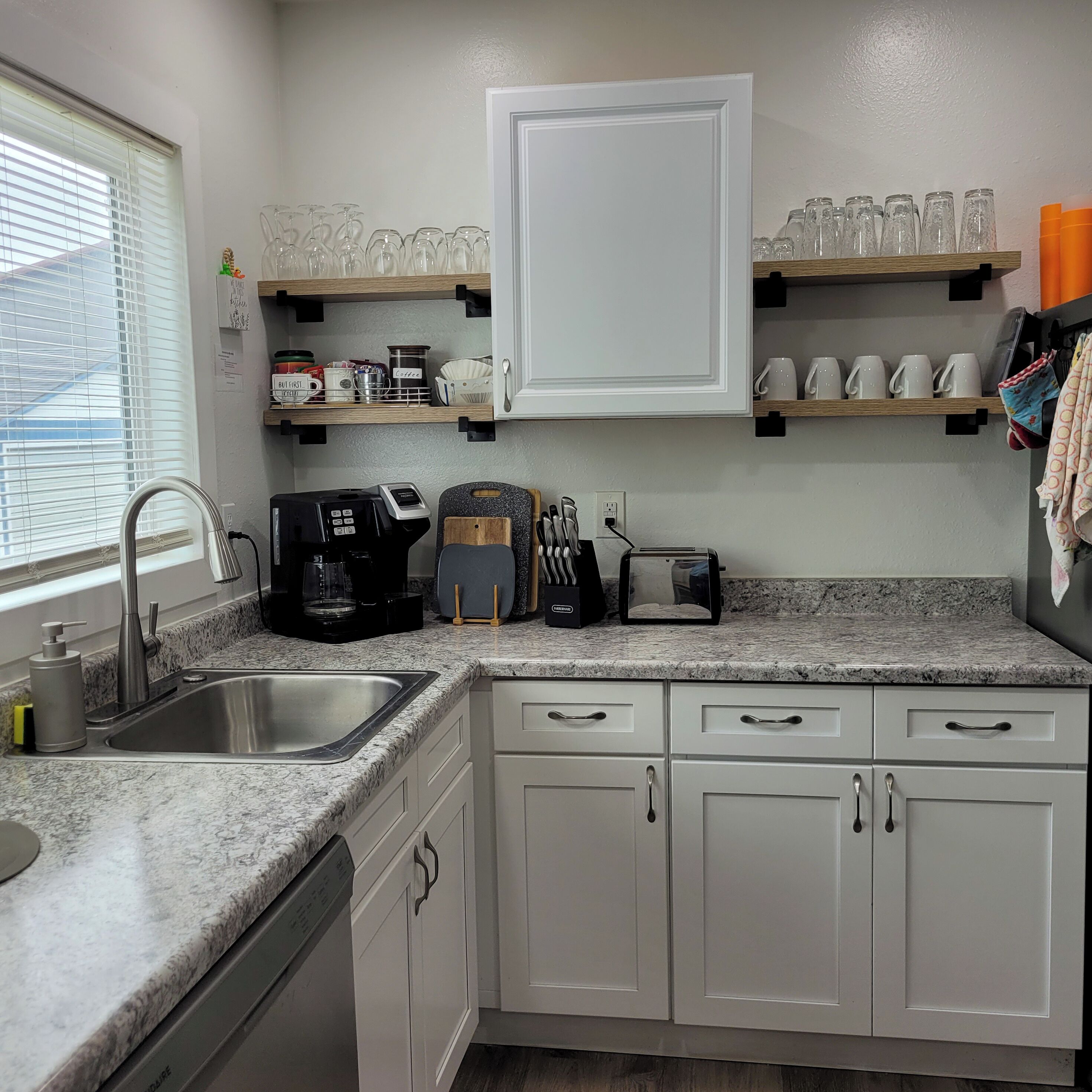 A kitchen with white cabinets, a granite countertop, a coffee maker, a toaster, and shelves above holding glassware and mugs.