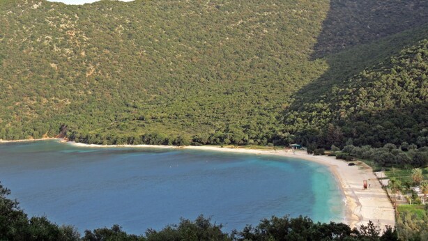 Paisagem costeira com uma praia, águas turquesa cristalinas e um morro arborizado.