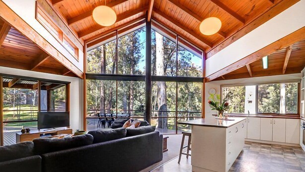 A spacious living area with a large sofa, a kitchen with white cabinets, and large windows offering a view of the outdoors. - Manna Gum - Base of Buller (Sawmill Settlement)