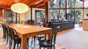 A spacious dining area with a wooden table and chairs, large windows offering a view of the forest, and a cozy seating area with a sofa and cushions. - Manna Gum - Base of Buller (Sawmill Settlement)