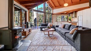 A spacious living area with a large sofa, a coffee table, and a wood-burning stove. There are large windows providing natural light and views of the outdoors. - Manna Gum - Base of Buller (Sawmill Settlement)