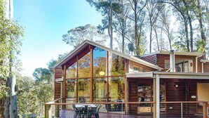 A modern wooden cabin with large glass windows, a deck with outdoor seating, and a satellite dish. - Manna Gum - Base of Buller (Sawmill Settlement)