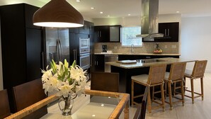 A modern kitchen with dark cabinetry, a central island, and a dining area with wicker chairs and a glass table. - Modern Abode 3 miles from beach  (Lake Worth Beach)