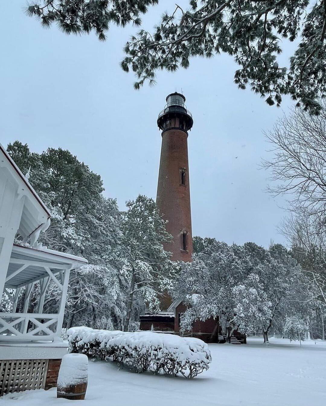 A snow-covered landscape with a lighthouse, a wooden structure, and snow-laden trees.