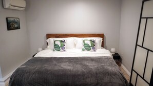 En snyggt bäddad säng med ett grått överkast och två kuddar med ett grönt och lila mönster. Det finns ett litet sängbord med en lampa, och en luftkonditioneringsenhet som är monterad på väggen.