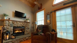 A cozy living room with a stone fireplace, a comfortable armchair, and a television mounted on the wall.