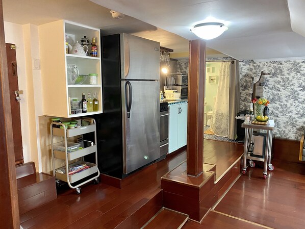 Una cocina con un refrigerador de acero inoxidable, un microondas y un lavabo. Hay un carro pequeño con frascos y botellas. El piso es de madera y las paredes tienen un estampado de papel tapiz floral.