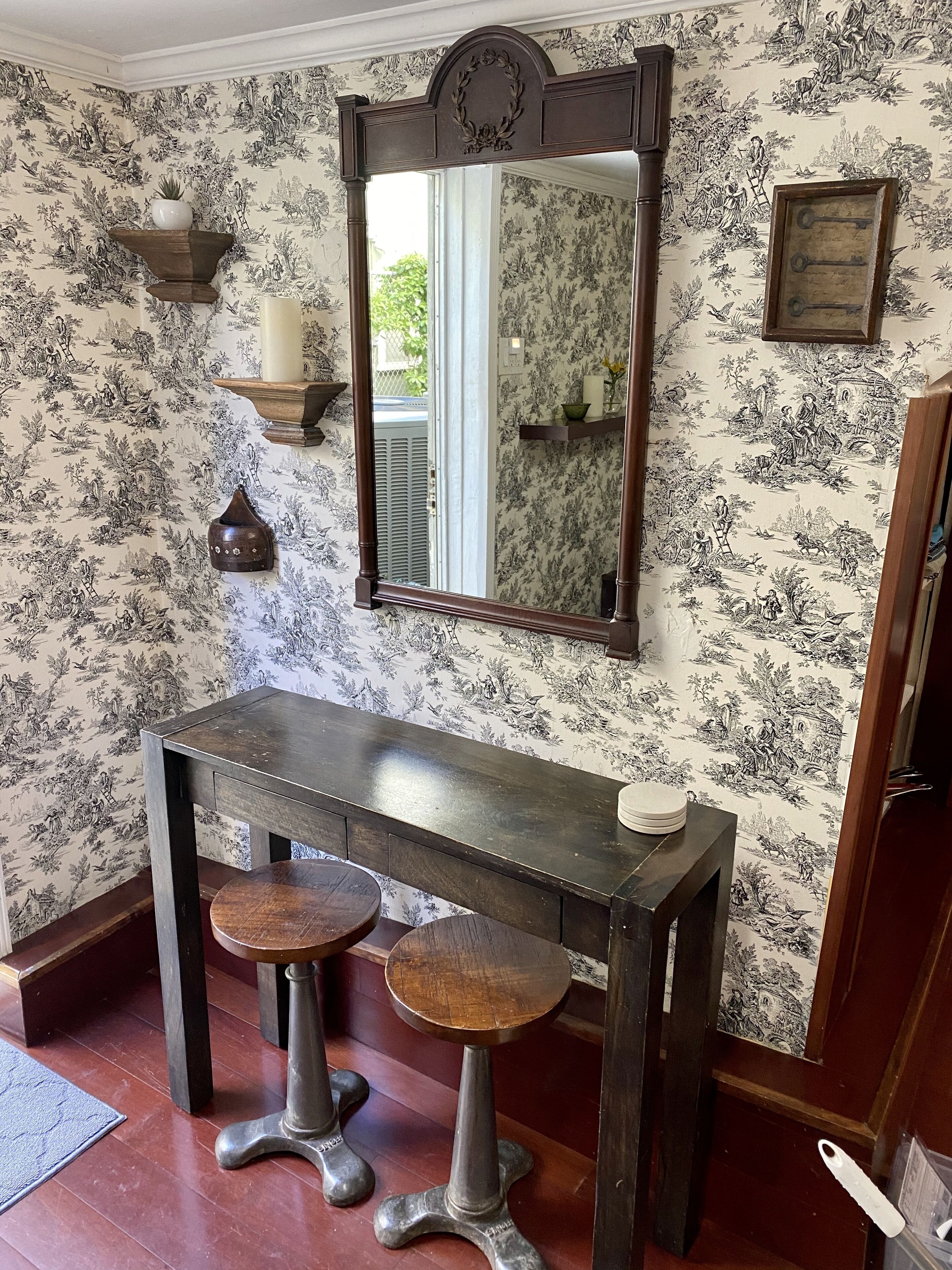 A room with a dark wooden table, two stools, a mirror, and wall-mounted shelves with decorative items.
