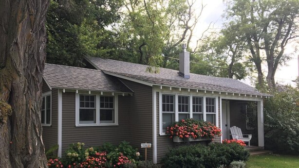 A single-story house with a gabled roof, a chimney, and a porch with a white Adirondack chair. There are flower boxes with red flowers on the porch, and a sign indicating the house number. - Apartment in the heart of Leland, just a short walk to Fishtown. (Leland)