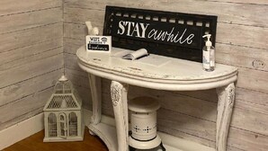 Un baño con un tocador blanco, un espejo con marco decorativo, un cartel que dice "STAY awhile", un farolillo blanco pequeño y una cita enmarcada en la pared.