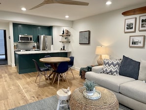 A modern kitchen with teal cabinets, a dining area with a round table and blue chairs, a living room with a beige sofa and a lamp, and a wooden floor.