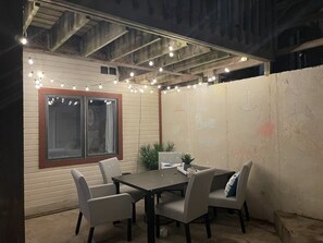 Un patio al aire libre con mesa y sillas, luces colgantes y una ventana con cortinas.