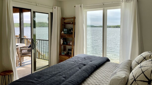 A bedroom with a bed, a wooden shelf, and a view of a lake through large windows. - Rosies place (Kimberling City)