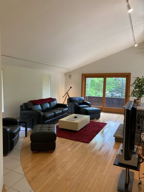 Ein modernes Wohnzimmer mit einem schwarzen Ledersofa, einem weißen Couchtisch und einem roten Teppich. Ein großes Fenster bietet Blick auf viel Grün draußen.
