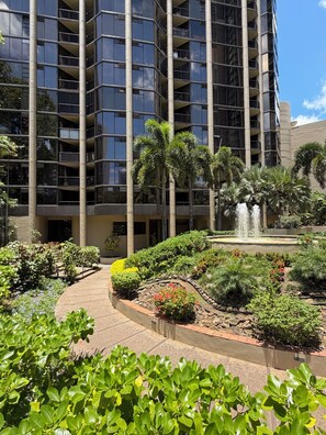 Um edifício com vários andares e um pátio central com uma fonte, rodeado por vegetação exuberante e palmeiras.
