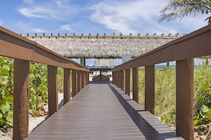 Un paseo marítimo de madera que conduce a una estructura con techo de paja, con vistas a una masa de agua y edificios a lo lejos.